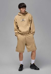 Młody mężczyzna w beżowej bluzie z kapturem Jordan i szortach, stojący z rękami w kieszeniach, patrzący w bok na jednolitym szarym tle.