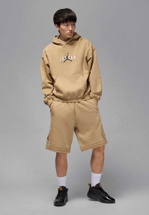 Giovane uomo con felpa con cappuccio e pantaloncini Jordan color cammello, in piedi con le mani nelle tasche, che guarda di lato contro uno sfondo grigio uniforme.