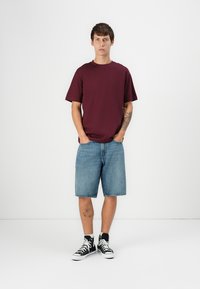 Burgunderfarbendes T-Shirt mit kurzen Ärmeln, hellblaue Jeansshorts und schwarze High-Top-Sneaker, stehend vor einem weißen Hintergrund. Schlichtes Design, lockere Passform.