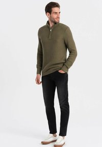 Olijfgroene textured breipullover met een half-rits kraag, gecombineerd met zwarte slim-fit jeans en witte sneakers met een tankleurige zool.