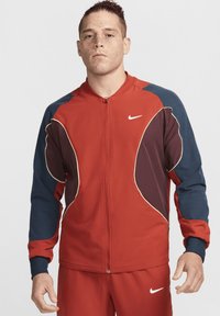Jachetă de sport roșie cu accente albastre și maron închis, cu închidere cu fermoar, manșete ribbed și logo-ul alb Nike pe piept.