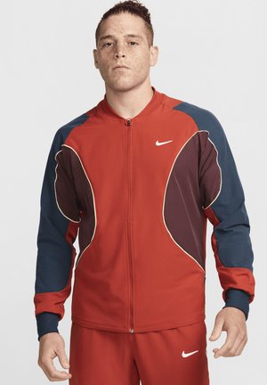 Veste de sport rouge avec des accents bleus et bordeaux foncé, arborant une fermeture éclair, des poignets côtelés et le logo blanc de Nike sur la poitrine.