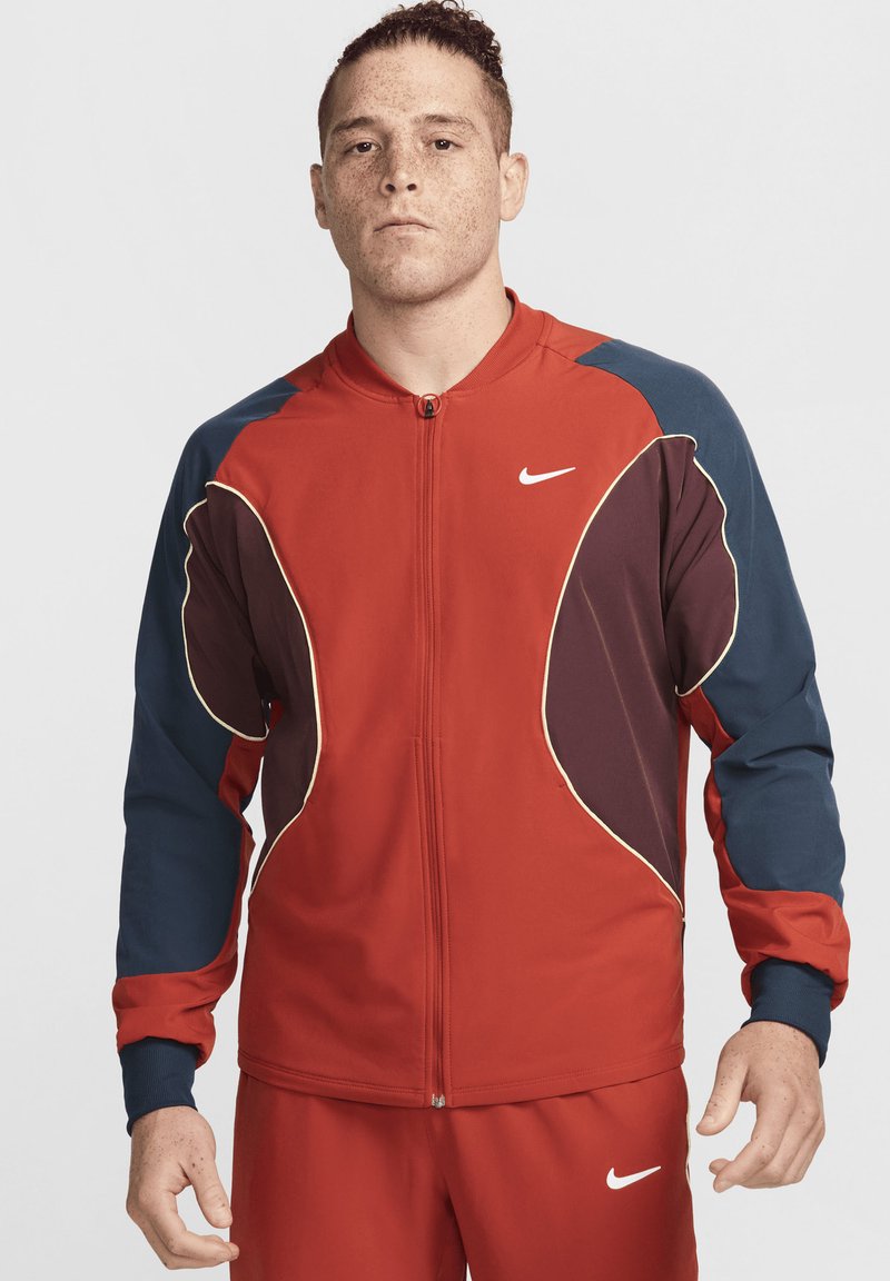 Jachetă de sport roșie cu accente albastre și maron închis, cu închidere cu fermoar, manșete ribbed și logo-ul alb Nike pe piept.