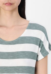 Lehký top s krátkým rukávem a kulatým výstřihem. Má střídající se zelené a bílé pruhy, hladkou texturu a volný střih.