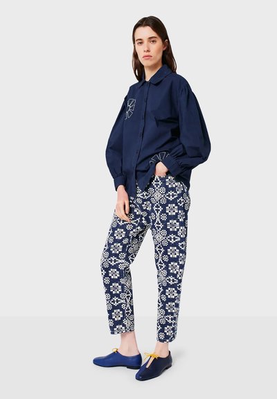 Donna in piedi che indossa una camicia azzurra con bottoni decorata con ricami, pantaloni a righe blu e bianchi, e scarpe blu con lacci gialli.