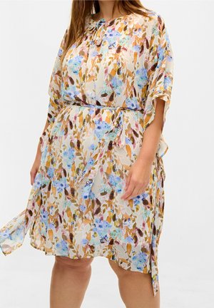 Femme portant une robe fleurie longueur genou avec des feuilles bleues, brunes et orange, dotée d'une ceinture nouée à la taille et de manches amples.