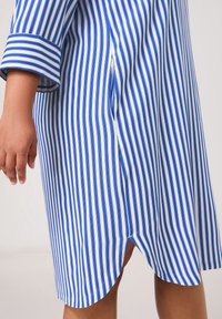 Robe chemise rayée présentant des rayures verticales bleues et blanches, un ourlet arrondi, des fentes latérales, et des manches trois-quarts. Texture de tissu lisse.