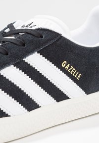 Basket en daim noir avec trois bandes en zigzag blanches, le mot "Gazelle" en lettres dorées et une semelle en caoutchouc blanc texturé.