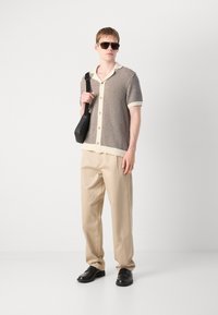 Short-sleeve knitted shirt with a textured, patterned design in brown and cream, paired with beige trousers and black shoes. Black shoulder bag.
