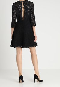 Robe en dentelle noire avec manches trois-quarts et jupe évasée. Caractérisée par un dos en V profond et un col festonné. Associée à des escarpins noirs à talons aiguilles.