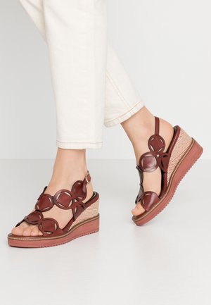 Pieds portant un pantalon blanc et des sandales compensées marron avec des brides en cuir à découpes circulaires, debout sur une surface unie gris clair.