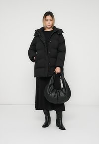 Schwarze Puffy-Jacke mit Kapuze, Reißverschluss und Druckknöpfen. Kombiniert mit einem langen schwarzen Kleid, schwarzen knielangen Stiefeln und einer schwarzen Handtasche.