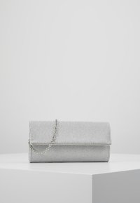 Pochette pailletée argentée avec rabat et fine chaîne argentée, présentée sur un piédestal blanc devant un fond gris clair.