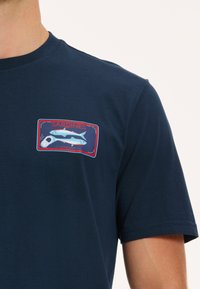 Tamsiai mėlyna medvilninė marškinėliai su grafiniu atvaizdu, kuriame pavaizduoti du šviesiai mėlyni žuvys ir skardinė, pažymėta "SARDINES" raudonomis ir mėlynomis akcentais.