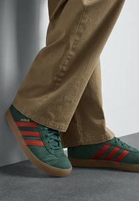 Zelené semišové tenisky s červenými pruhy a gumovou podrážkou, nošené s béžovými kalhotami s širokými nohavicemi, částečně viditelné na šedém pozadí.