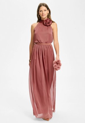 Frau trägt ein ärmelloses dusty-rose Halterneck-Kleid mit Blütendetail auf der Schulter, hält einen passenden Blumenstrauß und steht vor weißem Hintergrund.
