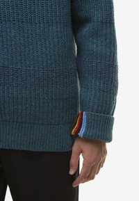 Πλεκτό πουλόβερ σε τιρκουάζ με υφασμάτινο μοτίβο, με ένα πολύχρωμο ριγέ τελείωμα στον καρπό. Ορατό χέρι στο μανίκι.