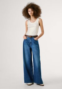 Jeans in denim blu a gamba larga abbinati a una canotta aderente color crema e una cintura marrone con accenti blu. Design semplice, texture liscia.