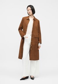 Lühikeste mustade juustega naine, kes kannab pruuni nööpidega pikka mantlit, valget topi, valgeid laiu pükse ja musti teravnurkseid kingi, seistes valge tausta ees.