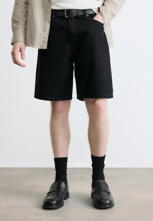 Personne portant un short noir jusqu'aux genoux, une ceinture noire, des chaussettes noires, des chaussures habillées noires, une chemise beige à rayures avec une main dans la poche.