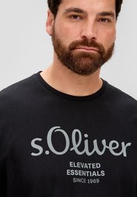 Must mustvalge särk, millel on hall tekst brändilt "s.Oliver", allpool on kirjas "ELEVATED ESSENTIALS" ja "SINCE 1969". Lühikesed varrukad.