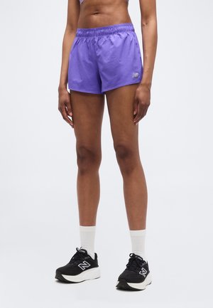 Persoon die paarse sportshorts draagt, zwarte en witte New Balance hardloopschoenen, en witte enkelsokken, staand tegen een effen achtergrond.