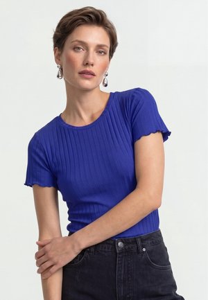 Frau mit kurzen braunen Haaren, die ein blaues geripptes Kurzarmtop und dunkle Jeans trägt, posiert mit verschränkten Armen vor einem hellen Hintergrund.