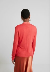 Pull en corail avec un dos texturé et des poignets côtelés, associé à une jupe en satin rouille, vue de l'arrière.