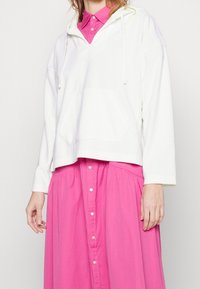 Bílá zkrácená mikina s kapucí a přední kapsou, přeložená přes růžové košilové šaty. Tkanina vypadá měkce a má mírnou texturu.