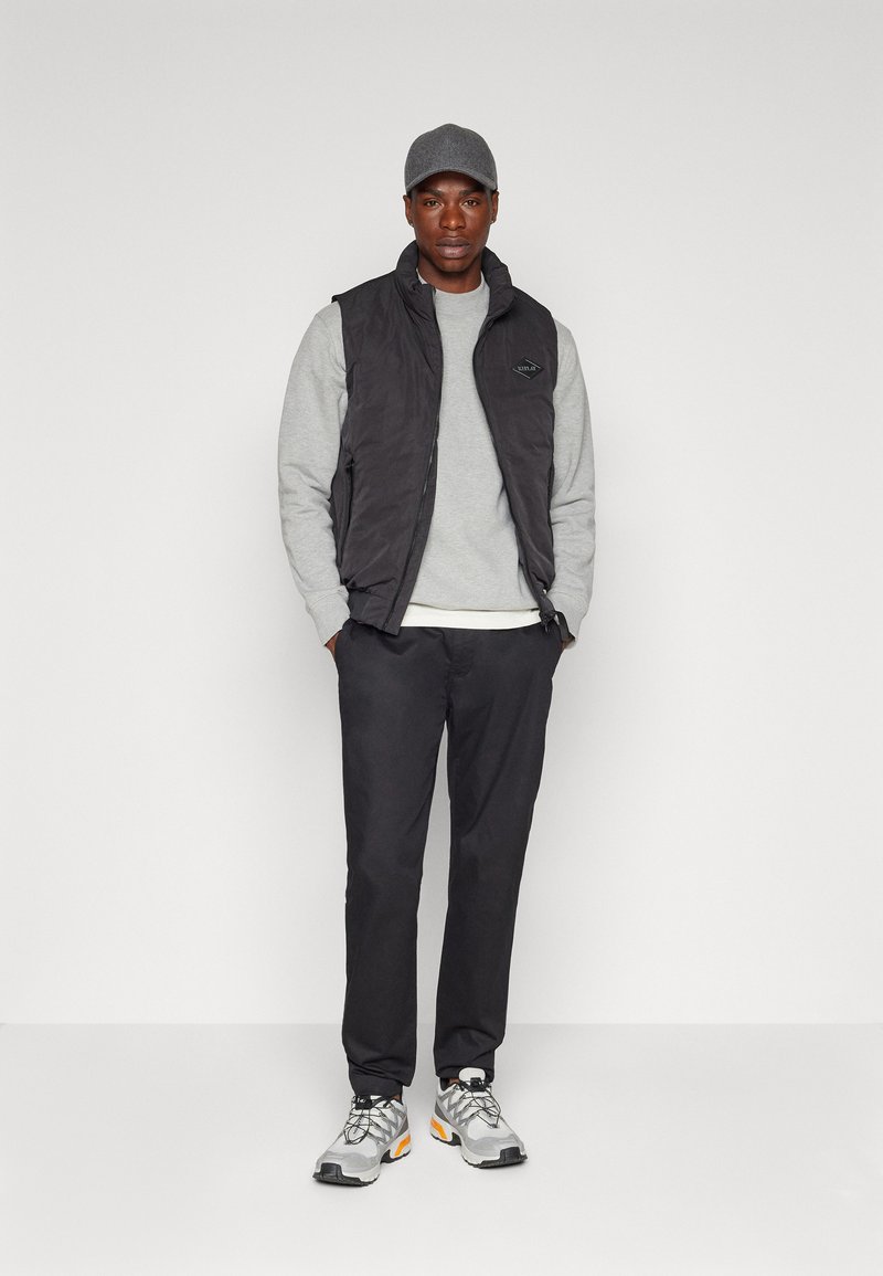 Schwarze gepolsterte Weste über einem grauen Sweatshirt, kombiniert mit schwarzen Hosen und grauen Sneakers mit orangefarbenen Akzenten. Das Model trägt eine graue Mütze.