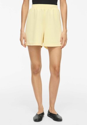 Personne porte un short jaune pâle à taille élastique, un haut noir et des chaussures plates noires, debout sur fond blanc.