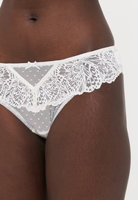Thong alb din dantela, cu plasă cu buline, margine dantelată ondulată și un accent cu fundiță mică în față. Material moale și ușor.
