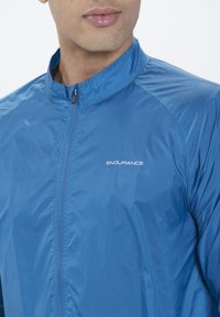 Blå lättviktsjacka med hög krage, dragkedjestängning och strukturerad tyg. Har en logotyp på framsidan och en figurnära design.