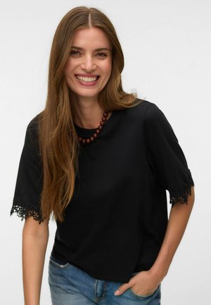 Femme souriante aux longs cheveux bruns portant un top noir à manches courtes avec bordure en dentelle et un jean bleu, main dans la poche, et un collier marron en perles.