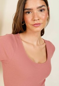 Kumerdatud, lühikeste varrukatega pluus pehmes roosat värvis laia ümmarguse kaelusega. Kangas näeb välja veniv ja tekstureeritud. Mudel kannab kuldseid ringkõrvarõngaid.