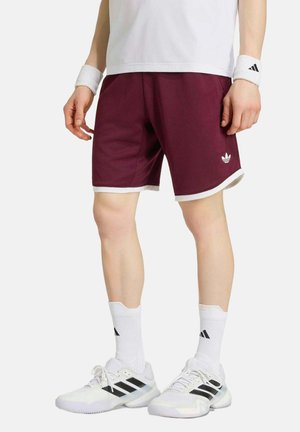 Persoon draagt donkerrode Adidas short, witte sportsokken, witte Adidas sneakers, witte polsbandjes en een wit shirt tegen een effen achtergrond.