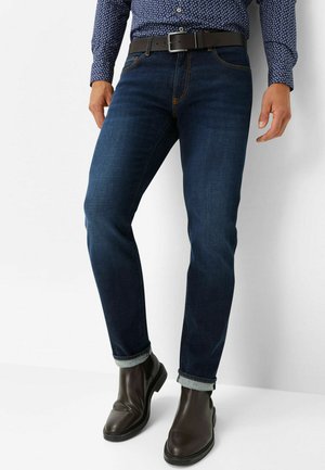 Dunkelblaue Denim-Jeans mit schmaler Passform, aufgerollten Säumen und kontrastierendem Stitching; kombiniert mit schwarzen Leder-Stiefeletten und einem gemusterten Hemd.