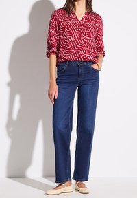 Kobieta ubrana w czerwoną bluzkę z długim rękawem w wzory, ciemnoniebieskie jeansy o prostych nogawkach, beżowe baleriny oraz złota bransoletka, stojąca przy białej ścianie.