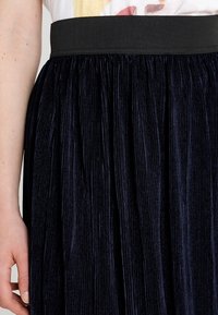 Falda plisada en azul marino con una ancha cinturilla negra. La tela tiene una textura suave con líneas verticales que crean una apariencia estructurada.