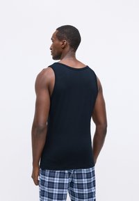 Μαύρο βαμβακερό αμάνικο μπλουζάκι με στρογγυλή λαιμόκοψη, φορώντας ένα μοντέλο που φοράει καρό σορτσάκια, δείχνοντας άνετη εφαρμογή και λεία υφή.