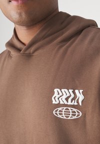 Hanorac maro cu un logo texturat alb "BRLN" și un design cu glob pe piept. Material moale, cu o croială lejeră și glugă.