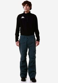 Pullover in pile nero con zip a metà e logo bianco; pantaloni da neve impermeabili verdi scuri con più tasche; robusti stivali neri.