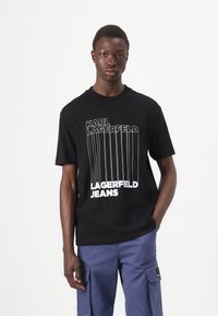 Svart bomull t-skjorte med hvit vertikal tekst som leser "KARL LAGERFELD" og "LAGERFELD JEANS." Korte ermer og rund halsdesign.