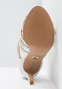Sandale blanche à talons hauts avec un design à brides, semelle texturée visible et petit logo en métal près du talon, taille 37.