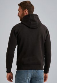 Zwarte hoodie met een soepele textuur en geribde manchetten. Voorzien van een kangoeroezak, gedragen met lichtblauwe jeans.