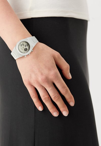 White rubber watch with a round face, silver accents, and three chronograph dials; worn on a hand resting against a black garment.