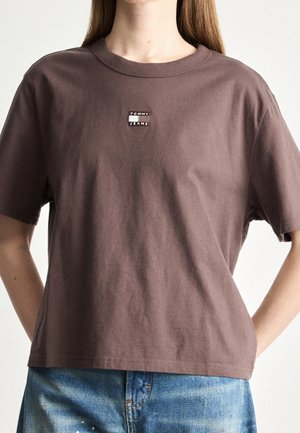 Vrouw die een bruin Tommy Jeans T-shirt met korte mouwen en een blauwe spijkerbroek draagt, staand met haar handen achter haar rug.