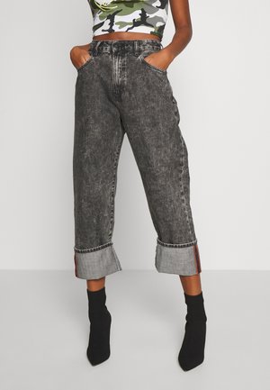 Personne portant un jean noir délavé taille haute avec revers, les mains dans les poches, des bottines chaussettes noires, associée à un crop top camouflage.