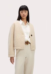 Beige teddyjakke med en kantet pasform, der har en enkelt sort knap og vide ærmer, båret over en hvid skjorte og beige bukser.