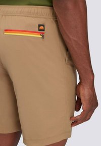 Pantaloni corti marrone con una finitura testurizzata, dotati di una tasca con zip rossa, gialla e arancione e un'etichetta con logo vicino alla vita.